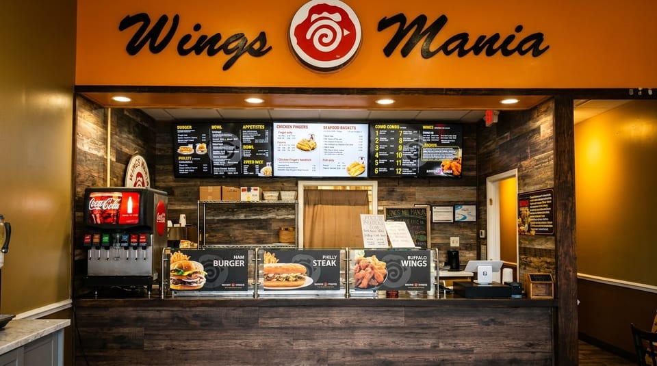The counter of a Wings Mania restaurant, featuring menus, a soda fountain, and food displays.