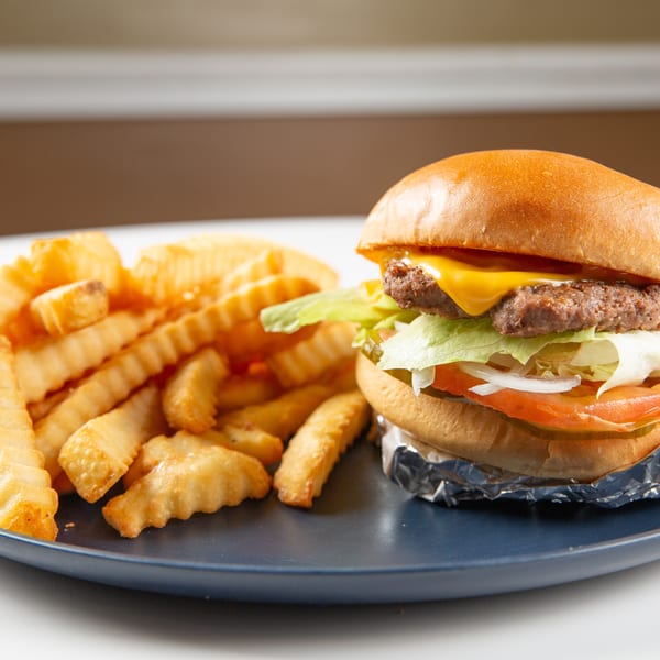 Cheeseburger with lettuce, tomato, onion, and pickles, wrapped in foil, next to crinkle-cut fries on a blue plate.