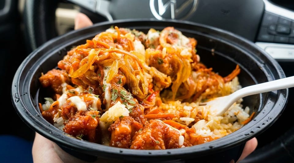 A person holds a black bowl of Korean fried chicken, rice, and kimchi with a white fork.
